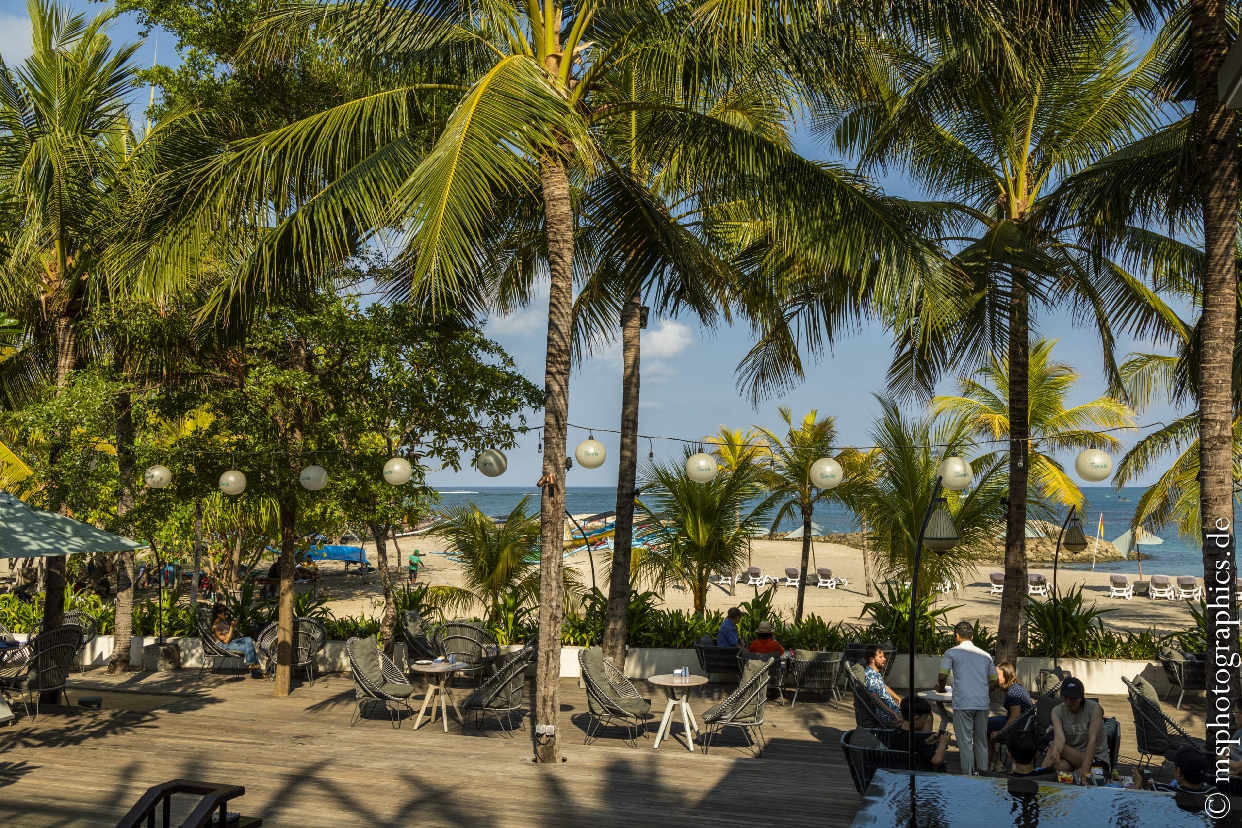 Tropische Palmen umgeben eine gemütliche Terrasse mit Sitzgelegenheiten und Blick auf den sandigen Strand und das Meer.