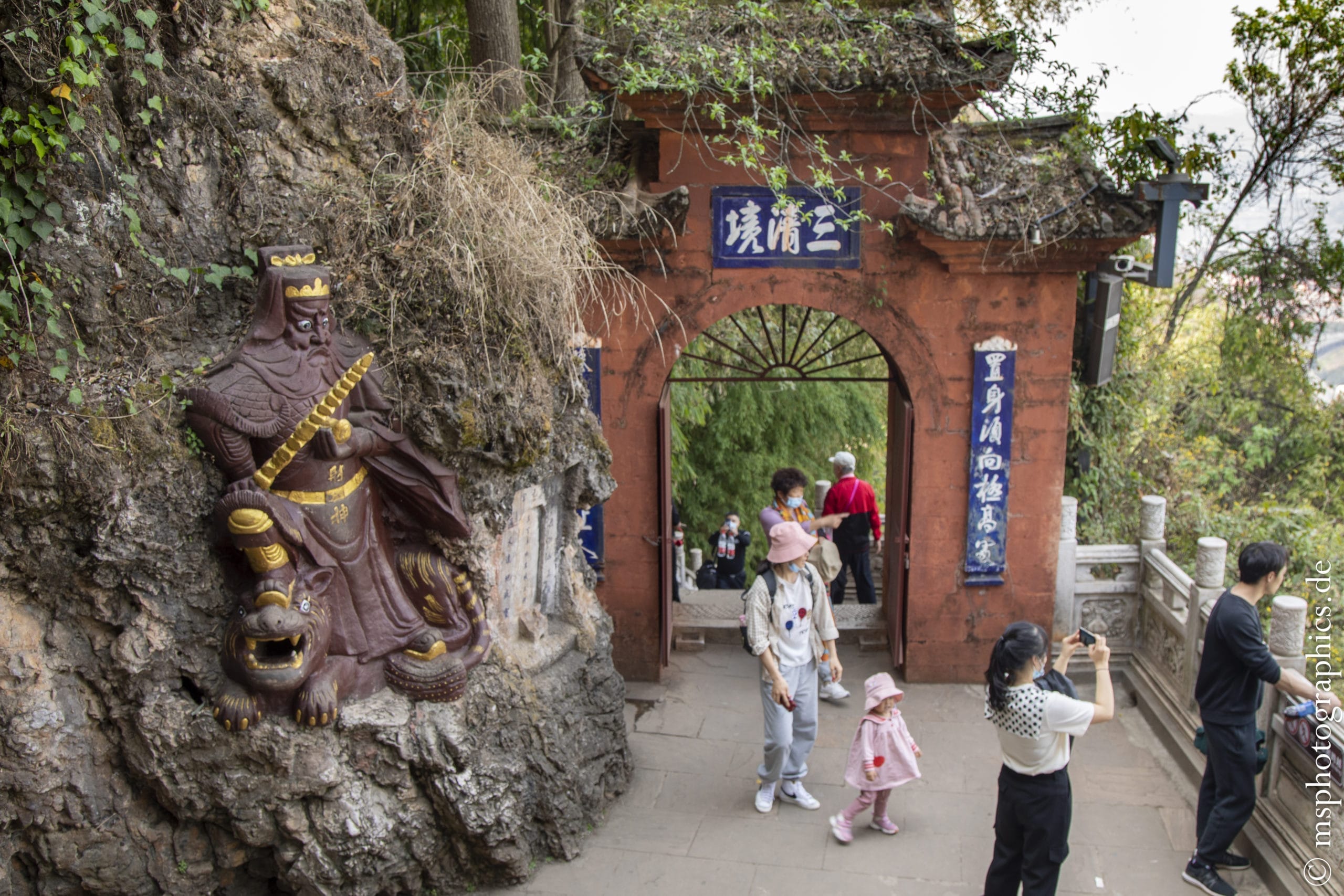 Eine Gruppe von Menschen in farbiger Kleidung steht vor einem Tor mit chinesischen Schriftzeichen, während eine Statue eines Kriegers an einem Felsen lehnt.